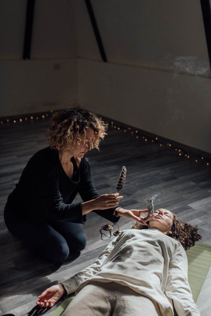 A Woman Holding a Feather and White Sage Beside a Woman Lying on Floor